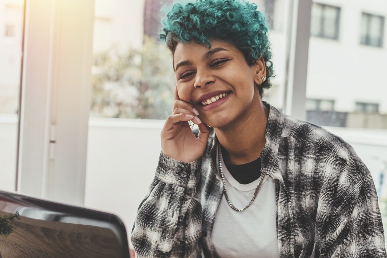 A student sits facing us, smiling at a computer. They have short green curly hair and are wearing a plaid shirt.