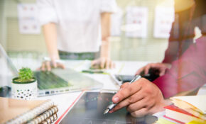 A closeup photo of a hand holding an old fashioned pen with a metal nib surrounded by books notebooks and a laptop