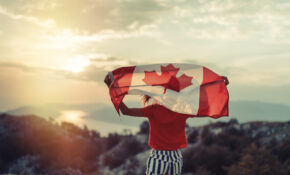 A person holding a Canadian flag aloft so it drapes them, as they look into the sunrise. Their back is to the camera.