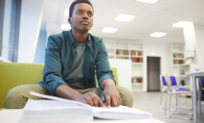 A student sits in a green chair with a book open in front of him on a low table. He is looking up and away, deep in thought.