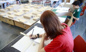 A student seated at a table, viewed from the back. They are wearing a red shirt and sketching a design.