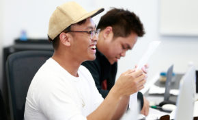 Two students are in a classroom. One is wearing a yellow baseball cap and laughing, the other is looking down at their work.