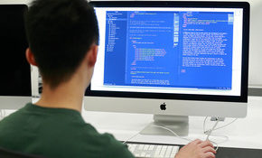 A student, seen from behind, sits at a monitor and works on code.