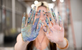 A student with bright blonde hair faces the camera holding her paint-covered hands up, obscuring her face.