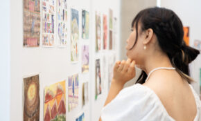 A student examines artworks on a wall. She is wearing a flowy white top and pearl jewelry.