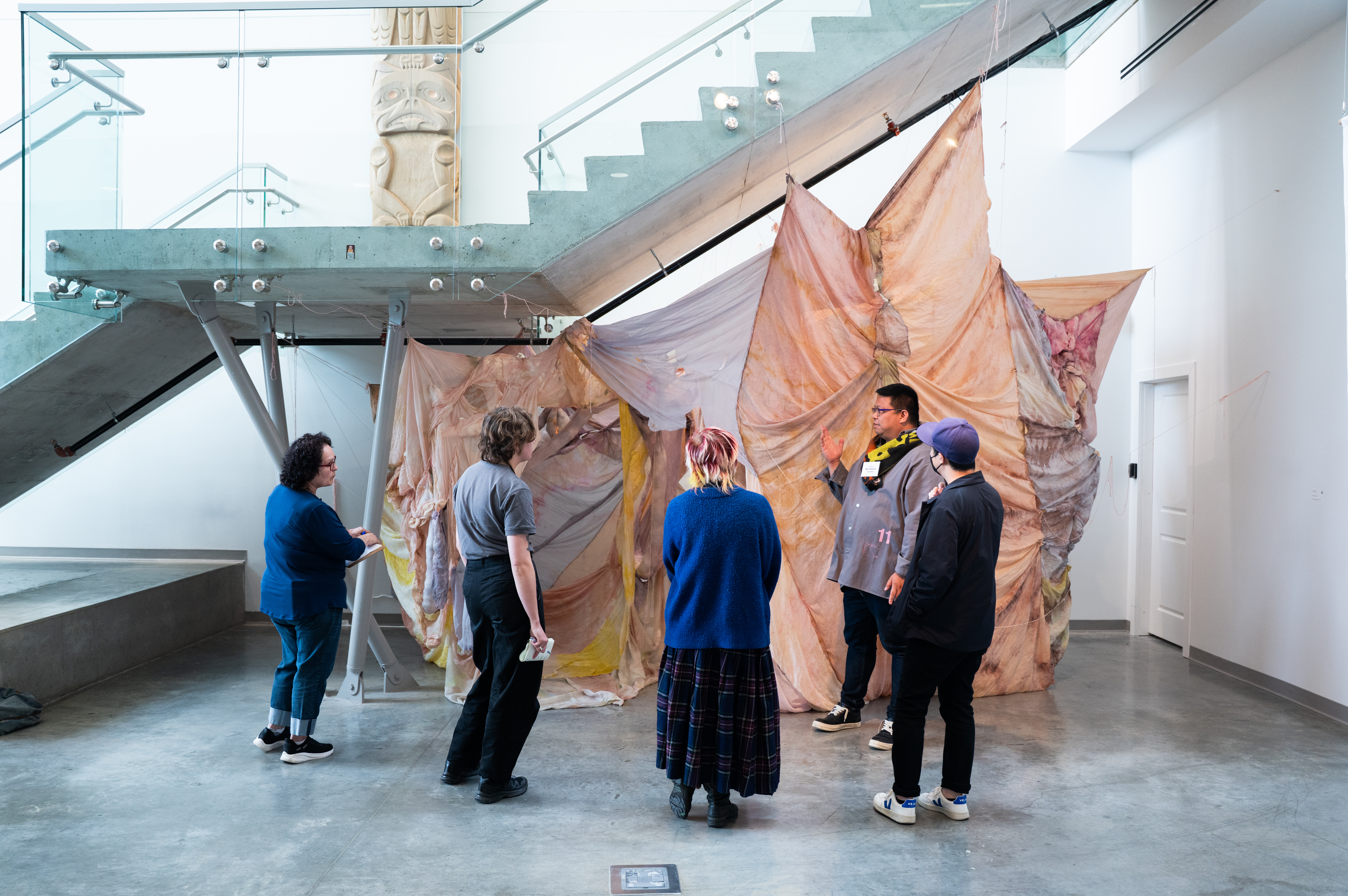 A group of five stand under stairs and in front of a fabric artwork piece