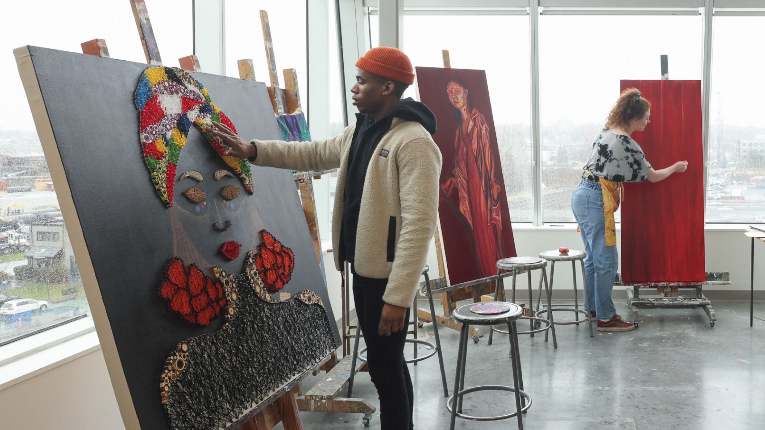 A student wearing a red hat works on a painting in ECU's printmaking studio. Another student is in the background, also working on a painting.