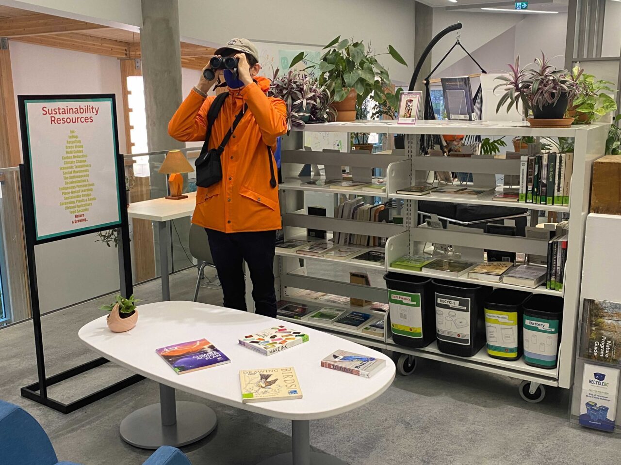 The ECU Library's Sustainability Resources Centre. A man in a orange coat holding a set of binoculars to his face in front of a book case featuring books and plants.
