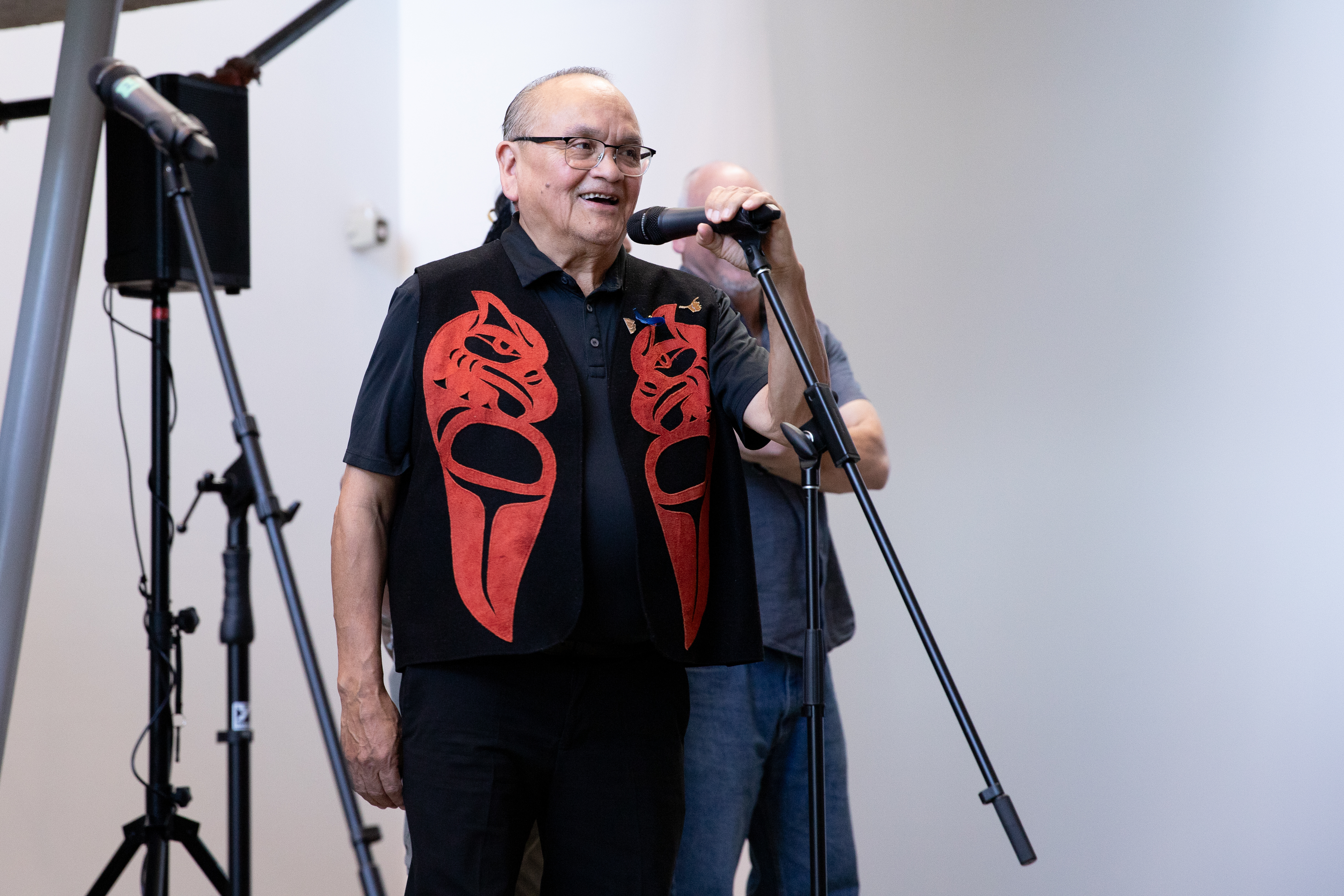 A man in a red and black waistcoat with Indigenous detailing speaks in front of a microphone
