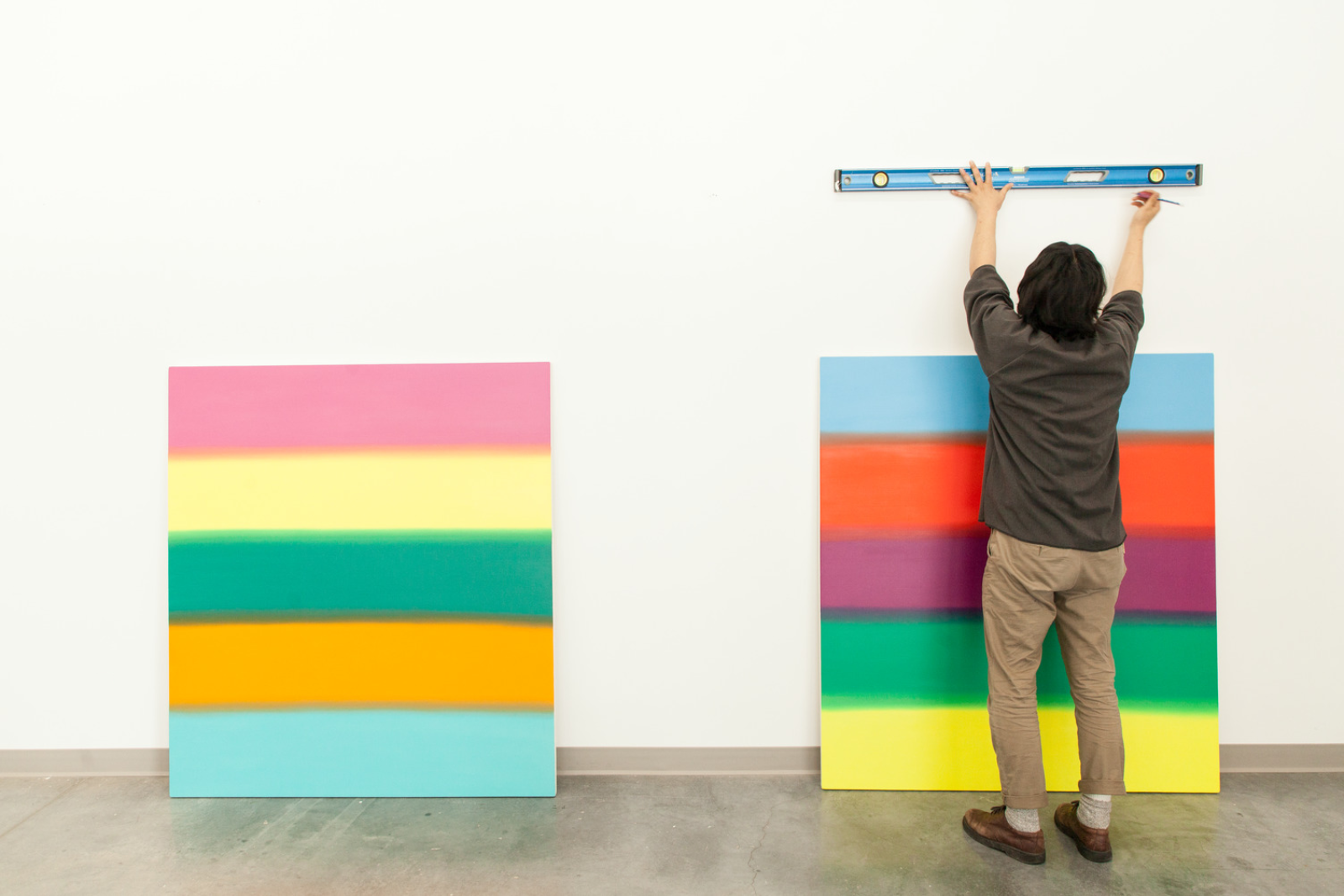 A student with their back to the camera holds a level to the wall, in the midst of hanging two colourfully painted canvases.