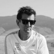 Black and white photo on white background of instructor with short dark hair wearing a white t-shirt and sunglasses.