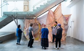 A group of five stand under stairs and in front of a fabric artwork piece