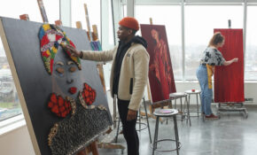 A student wearing a red hat works on a painting in ECU's printmaking studio. Another student is in the background, also working on a painting.