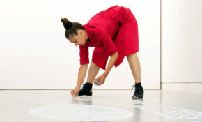 A student in a red dress bends down to add sand to a design on the floor.