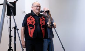 A man in a red and black waistcoat with Indigenous detailing speaks in front of a microphone