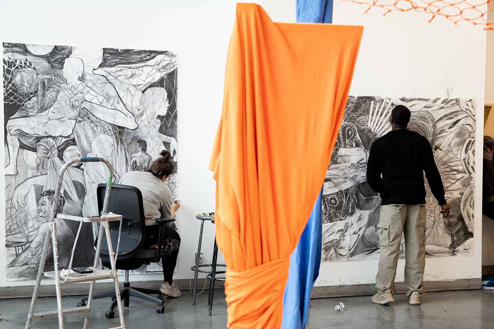 Two people, divided by an orange textile in the foreground, work side-by-side in a studio on large-scale drawings.