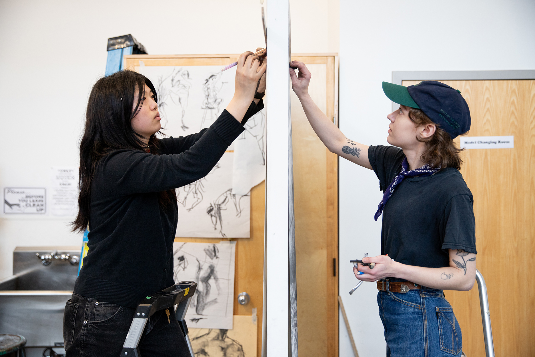 Two people work on separate drawings hung on either side of a room divider.