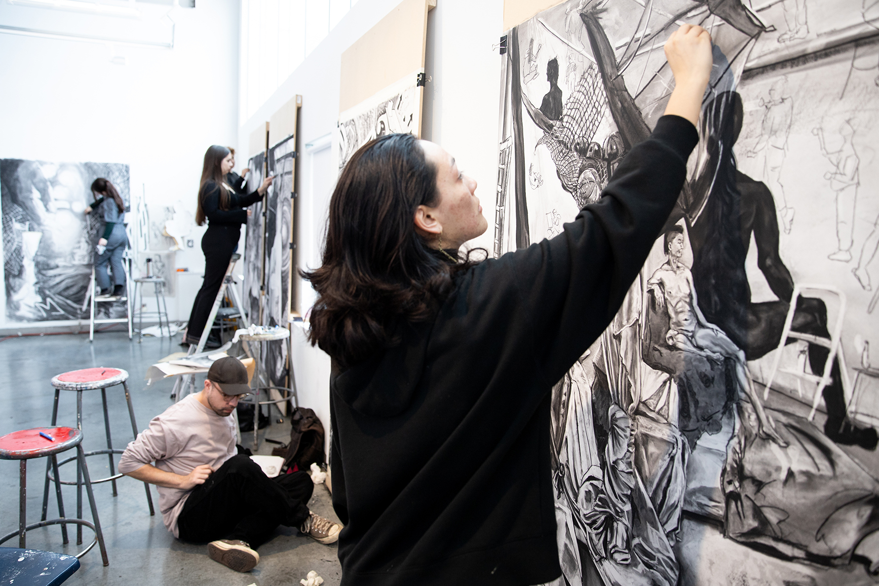 A group of students in a large studio work at the same time on separate, large-scale drawings.