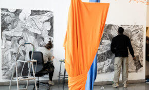 Two people, divided by an orange textile in the foreground, work side-by-side in a studio on large-scale drawings.