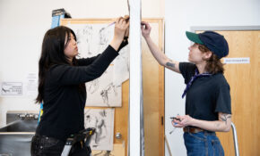 Two people work on separate drawings hung on either side of a room divider.