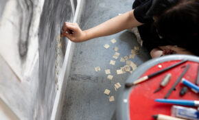 A person carefully applies small squares of collaged paper to a large black-and-white drawing.